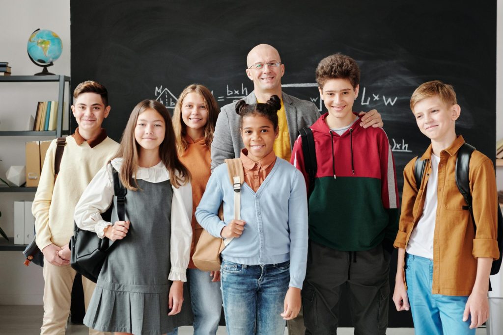 A group of diverse teenagers with backpacks and a teacher smiling in a classroom.
